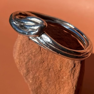May include: A silver-colored ring with a knot design. The ring is resting on a reddish-brown stone against a matching background. The ring's design features a knot at the top, with a smooth, polished finish.