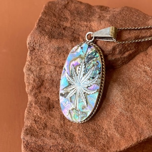 Puede incluir: Un collar de cadena de plata con un colgante ovalado que presenta una incrustación detallada de concha de abulón de una hoja de marihuana. El colgante tiene un borde de plata y es iridiscente con tonos de azul, verde y morado.