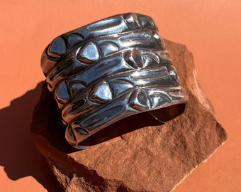 May include: A silver metal cuff bracelet with a textured design. The bracelet has a wide, flat band with a repeating pattern of raised circles and lines.