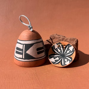 May include: Two small ceramic ornaments with geometric designs. The ornament on the left is bell-shaped and has a black and white geometric pattern. The ornament on the right is round and has a black and white floral pattern.