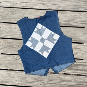 May include: A denim vest with a patchwork design on the back. The vest is a medium blue color and features a square patch with a white and blue star pattern. The vest is sleeveless and has a V-neck.