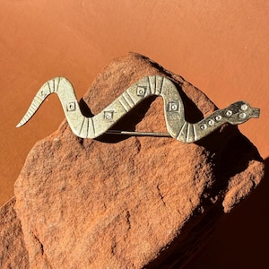 May include: A vintage-style snake brooch in a metallic gold color. The brooch is designed in a serpentine shape, with etched details and square accents along its body. It is resting on a reddish-brown stone, showcasing its intricate design.
