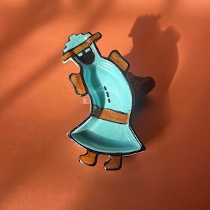 May include: A turquoise ceramic dish shaped like a person wearing a brown hat and brown shoes. The dish has a black outline and brown details.