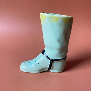 May include: A light blue ceramic boot-shaped vase with a black strap detail. The vase has a yellow glaze on the top edge and a few painted details on the side.