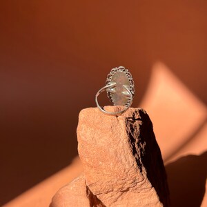 Op de afbeelding: Een zilveren ring met een grote ovale edelsteen gezet in een filigraan ontwerp. De ring ligt op een kleine steen.