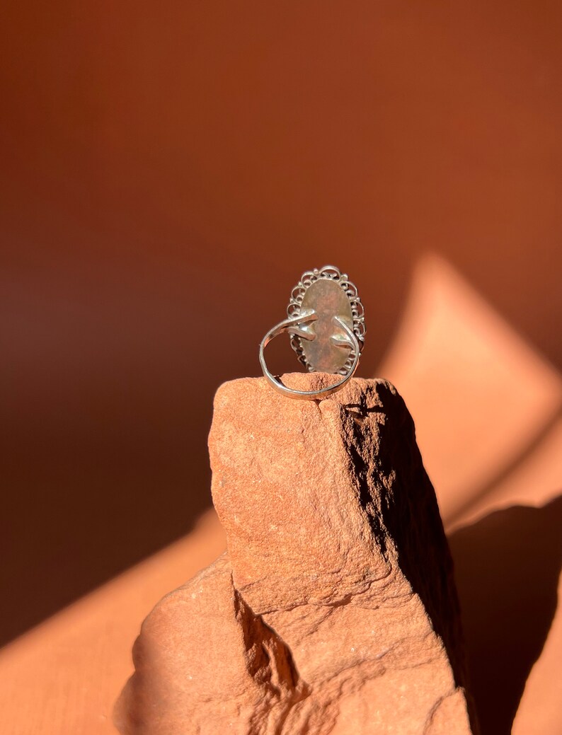 Op de afbeelding: Een zilveren ring met een grote ovale edelsteen gezet in een filigraan ontwerp. De ring ligt op een kleine steen.