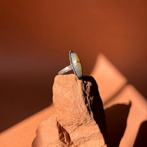 Op de afbeelding: Een zilveren ring met een ovale gele en witte steen gezet in een decoratieve zilveren band. De ring ligt op een kleine, ruwe, rode steen.