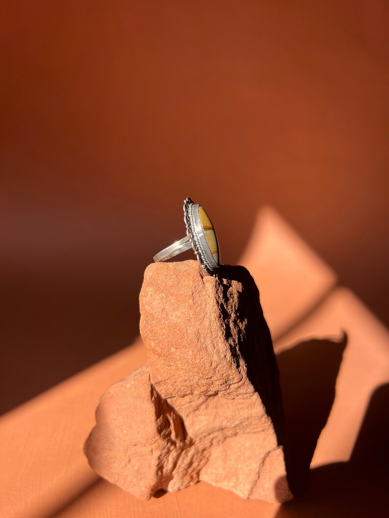 Op de afbeelding: Een zilveren ring met een ovale gele en witte steen gezet in een decoratieve zilveren band. De ring ligt op een kleine, ruwe, rode steen.