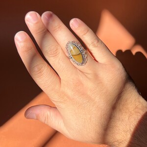 Op de afbeelding: Een zilveren ring met een ovale steen gezet in een filigraan ontwerp. De steen is een mix van bruine, gele en witte kleuren.