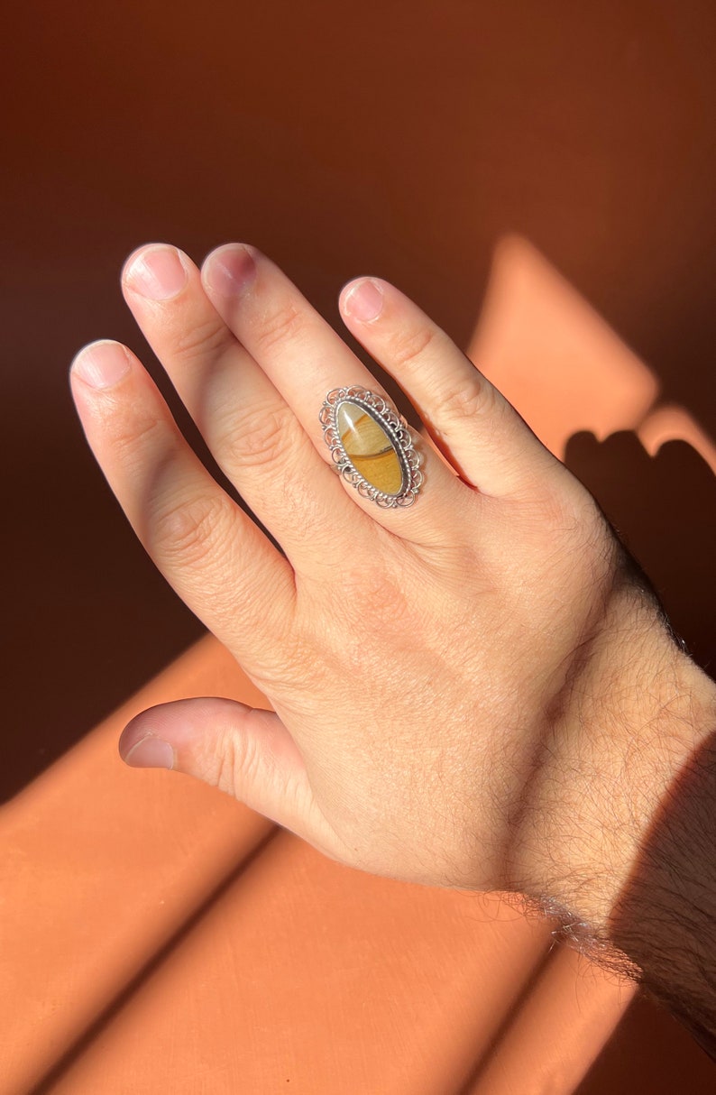 Op de afbeelding: Een zilveren ring met een ovale steen gezet in een filigraan ontwerp. De steen is een mix van bruine, gele en witte kleuren.