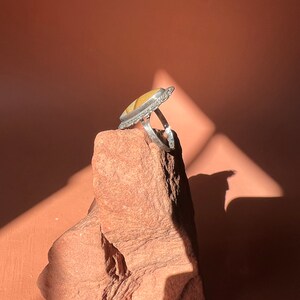 Op de afbeelding: Een zilveren ring met een gele en witte agaat-steen gezet in een zilveren bezel. De ring ligt op een rode rots.