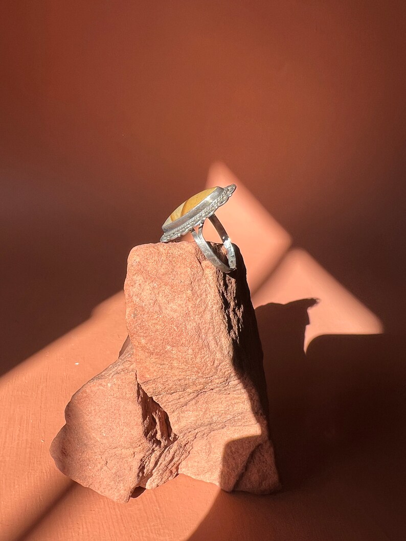 Op de afbeelding: Een zilveren ring met een gele en witte agaat-steen gezet in een zilveren bezel. De ring ligt op een rode rots.
