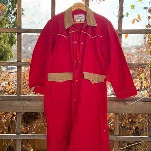 May include: A long, red coat with a tan corduroy collar and pocket accents. The coat has a button-up front and two front pockets. The coat is hanging on a wooden hanger. The coat is a vintage style.