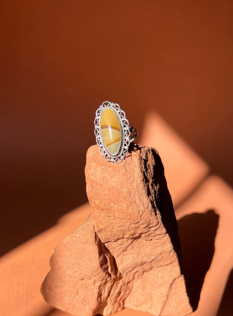 Op de afbeelding: Een zilveren ring met een ovale steen gezet in een filigraan ontwerp. De steen is een mix van bruine, gele en witte kleuren.
