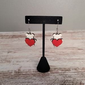 May include: A pair of dangle earrings featuring a white wooden bear silhouette holding a red heart. The earrings are hanging from a black display stand.
