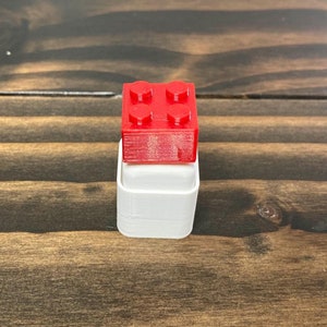 May include: A small red plastic toy brick sits on top of a white plastic container.
