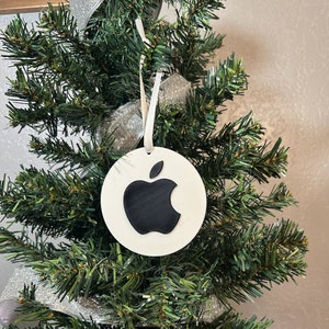May include: White round Christmas ornament with a black Apple logo. The ornament is hanging on a green artificial Christmas tree.