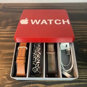 Może przedstawiać: Czerwone pudełko z logo Apple i napisem "WATCH" nadrukowanym na pokrywie. Pudełko jest otwarte i zawiera trzy paski do zegarka i kabel ładujący.