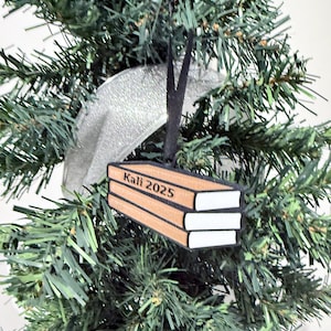 Puede incluir: Adorno navideño con forma de pila de libros. El adorno es marrón con páginas blancas y el texto "Kali 2025". Cuelga de una cinta negra, sobre un árbol de Navidad verde y una cinta plateada.