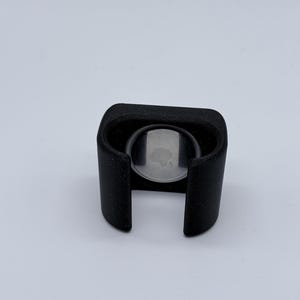 Könnte beinhalten: Ein schwarzer, 3D-gedruckter Halter für ein kleines, kreisförmiges Gerät. Der Halter hat ein U-förmiges Design mit abgerundeter Oberseite und einer kreisförmigen Öffnung zur Anzeige des Geräts. Das Gerät hat eine silberne Oberfläche mit einem Logo.