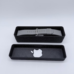 Puede incluir: Una correa de Apple Watch de cuero gris en una caja negra con un logotipo de Apple blanco.
