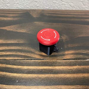 May include: A red push button with a black base and white arrows indicating direction. The button is on a wooden surface.