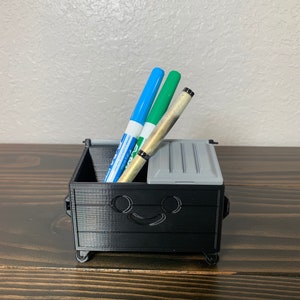 May include: A black 3D printed trash can with wheels, designed to look like a dumpster, with a smiling face. It is holding two blue and green markers and a gold pen.