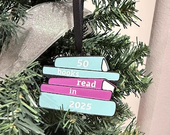 May include: A festive Christmas tree decoration featuring a stack of books. The ornament, in shades of light blue and purple, displays the text "50 books read in 2025". It's suspended by a black ribbon, set against a green Christmas tree and a silver ribbon.