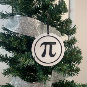 Puede incluir: Un adorno navideño circular blanco con un contorno negro y el símbolo matemático pi en negro en su interior. El adorno está colgado de un árbol de Navidad artificial verde con cinta de brillo plateado.