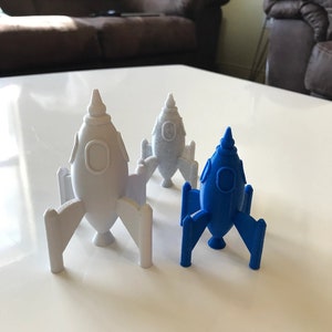 May include: Three toy rockets, two are white and one is blue. The rockets are standing on a white surface.
