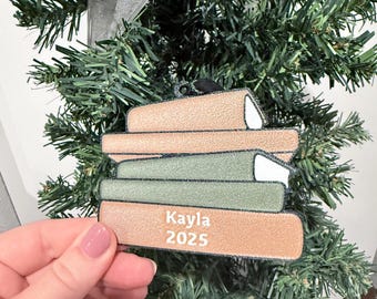 May include: A Christmas tree decoration in the shape of a stack of books. The ornament is tan and green, with the name "Kayla" and the year "2025" printed on the bottom book. A silver ribbon is attached for hanging.