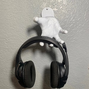 May include: A white 3D printed hand shaped headphone holder with a pair of black over-ear headphones hanging from it.