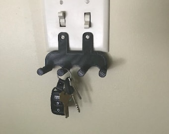 Puede incluir: Un porta llaves de plástico negro montado en una placa de interruptor de luz blanca con un juego de llaves colgando.