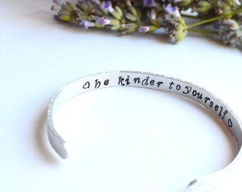 May include: Silver bangle bracelet with the inscription "Be kinder to yourself" and a small heart symbol.