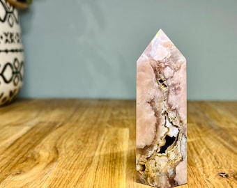May include: A pink and white crystal tower with a pointed top. The crystal has a marbled appearance with areas of light pink, white, and brown. The crystal is standing on a wooden surface.