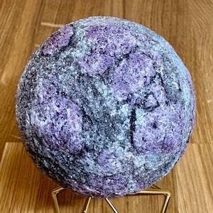May include: A spherical, textured stone in shades of purple and grey. The stone is presented on a gold-coloured geometric stand, highlighting its unique colour and pattern. The stone's surface has a rough, natural appearance.