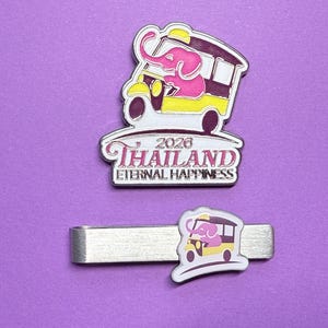 May include: A metal pin and a tie clip featuring a pink elephant riding in a yellow and dark purple tuk-tuk. The pin includes the text 2026 THAILAND ETERNAL HAPPINESS in pink and black lettering. Both items display the same colorful graphic design.