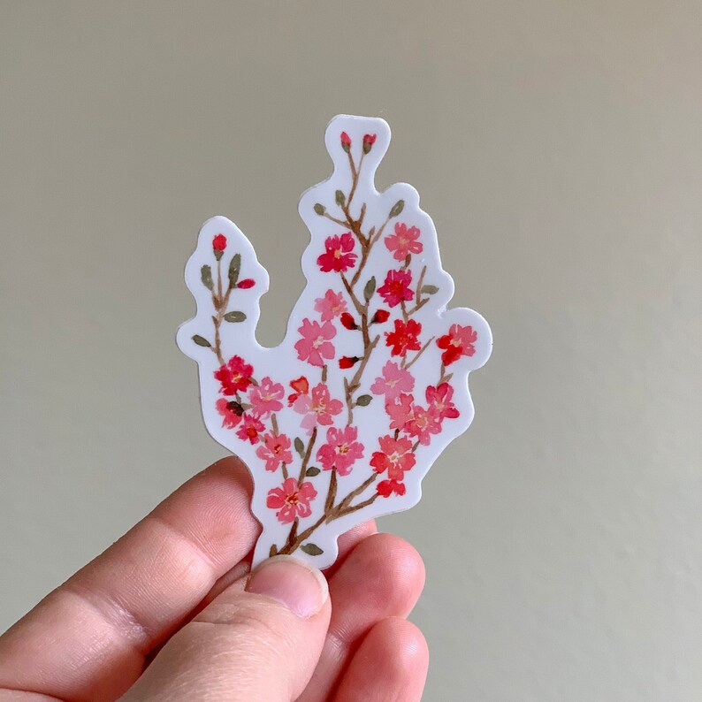 Sticker of a Sakura/ cherry blossom flower with green leaves and brown branches being held by model against a beige background. Sticker is professionally manufactured and features original watercolor artwork.