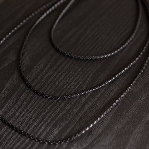 May include: A black braided cord necklace arranged in multiple circular loops. The cord features a consistent woven texture throughout its length, creating a uniform appearance. The material appears flexible and smooth, forming overlapping concentric circles.