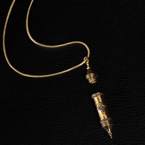 May include: A gold-toned necklace featuring a thin chain and a cylindrical pendant that is detached into two pieces. The pendant has a pointed base, a screw-top cap, and a small diamond-shaped decorative engraving on the main body section.
