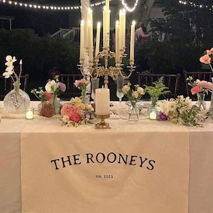 May include: A white table runner with black text that reads "THE ROONEYS est. 2023". A gold candelabra with white candles sits on the table runner, surrounded by floral arrangements.