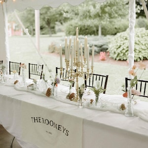 May include: A white table runner with the text "THE ROONEYS est. 2021" in black lettering. The table is set with white plates, clear glasses, and floral arrangements in glass vases. A gold candelabra with white candles is the centerpiece.
