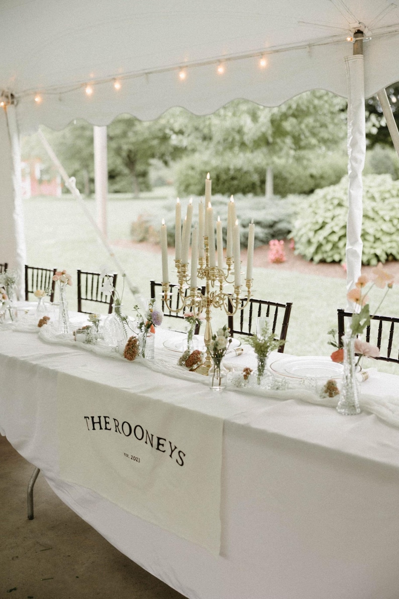 May include: A white table runner with the text "THE ROONEYS est. 2021" in black lettering. The table is set with white plates, clear glasses, and floral arrangements in glass vases. A gold candelabra with white candles is the centerpiece.