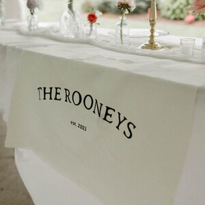 May include: White table runner with black text that reads "THE ROONEYS est. 2023".