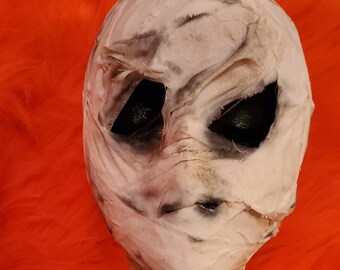 May include: A handmade Halloween mummy head sculpture. The head is wrapped in white fabric with dark shading and black eye sockets. The sculpture is set against a vibrant orange background.