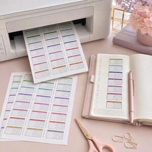 Puede incluir: Una impresora blanca imprimiendo hojas de pegatinas de calendario coloridas. Un cuaderno rosa con un bol&iacute;grafo, tijeras y clips est&aacute; sobre una superficie rosa claro. Las pegatinas de calendario tienen un dise&ntilde;o de cuadr&iacute;cula con cuadros de varios colores.
