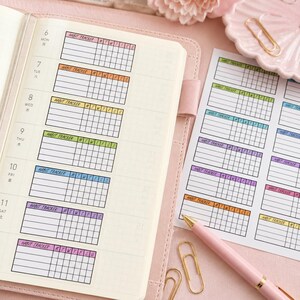 Puede incluir: Una agenda rosa abierta en una p&aacute;gina con pegatinas de seguimiento de h&aacute;bitos coloridas. Las pegatinas rectangulares tienen la inscripci&oacute;n "Habit Tracker" y casillas para el seguimiento de h&aacute;bitos diarios. Tambi&eacute;n se ve un bol&iacute;grafo rosa y clips dorados.