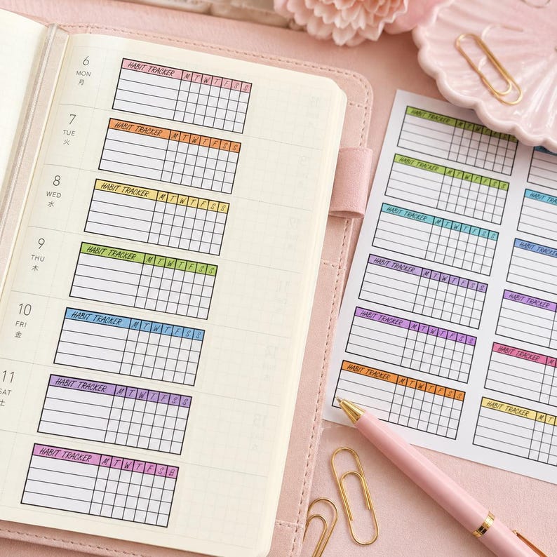 Puede incluir: Una agenda rosa abierta en una p&aacute;gina con pegatinas de seguimiento de h&aacute;bitos coloridas. Las pegatinas rectangulares tienen la inscripci&oacute;n "Habit Tracker" y casillas para el seguimiento de h&aacute;bitos diarios. Tambi&eacute;n se ve un bol&iacute;grafo rosa y clips dorados.