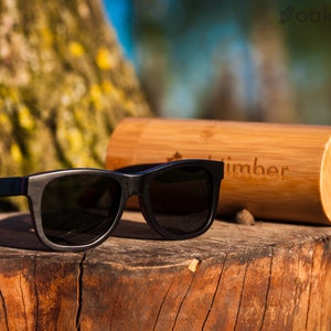 May include: Black sunglasses with a red accent on the side, resting on a wooden stump. The sunglasses are in a bamboo case with the word "oaktimber" engraved on it.
