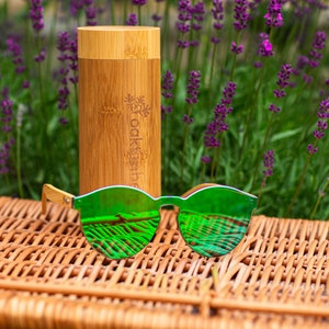 May include: Green mirrored sunglasses with a wooden frame, resting on a wicker surface. A bamboo case with the text "Oakley" is in the background.